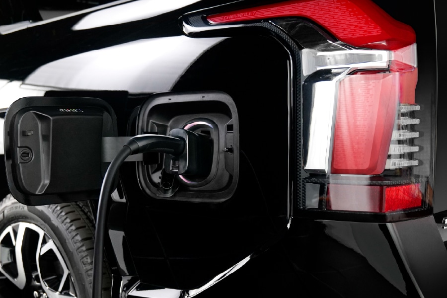 Close-up of an electric vehicle being charged. The charging cable is plugged into the port on the rear side of a black car. The cars taillight and part of the wheel are visible. The vehicle is shiny and sleek in design.