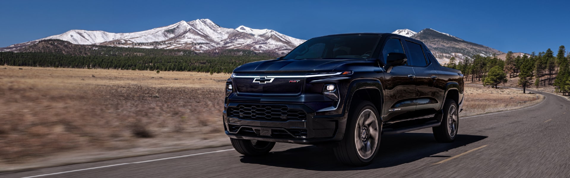 A black Chevrolet pickup truck drives on a paved road with snow-capped mountains and a clear blue sky in the background.