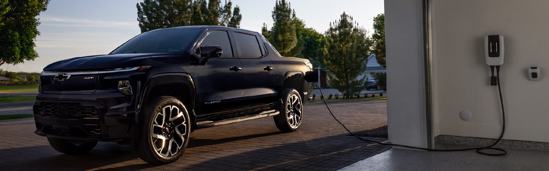 A black electric truck is parked in a driveway, connected to a charging station inside a garage. The scene is set against trees and a clear sky in the background, creating a calm and modern suburban environment.