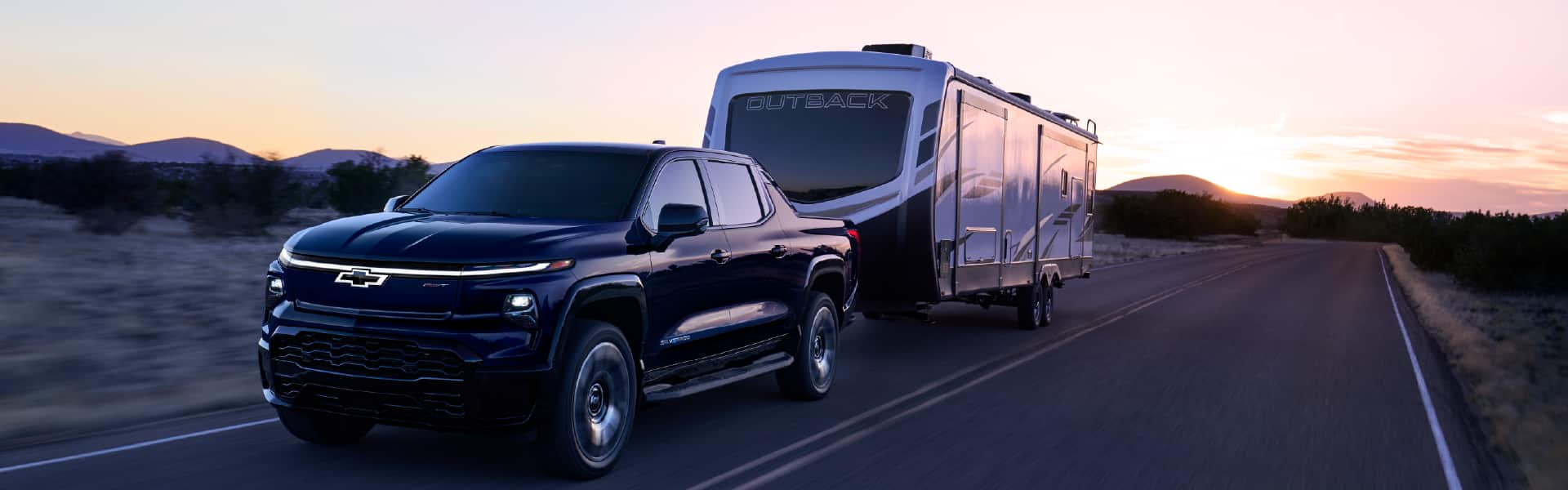 A dark blue pickup truck towing a large Outback trailer down an open road at sunset, with silhouettes of mountains in the background under a colorful sky.