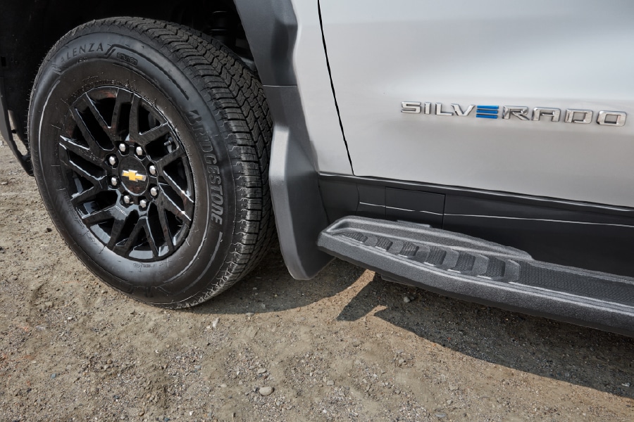 Close-up of a silver trucks side, showing a black wheel with the Chevrolet logo, a Bridgestone tire, and the SILVERADO emblem on the body. The ground is dirt, and theres a black step bar below the door.