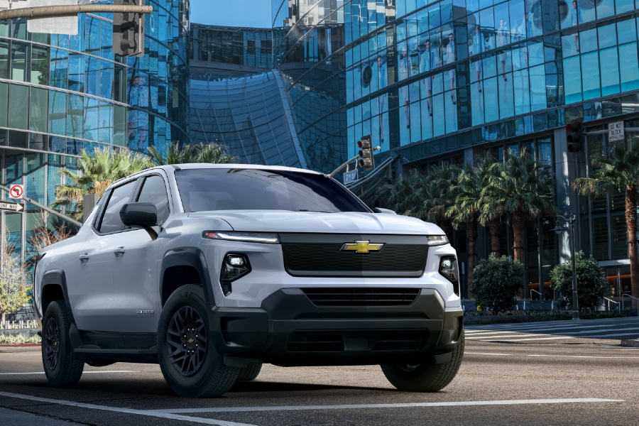 A white Chevrolet pickup truck is parked on a city street in front of a glass building. Palm trees and other skyscrapers are visible in the background. The trucks design is modern and sleek.