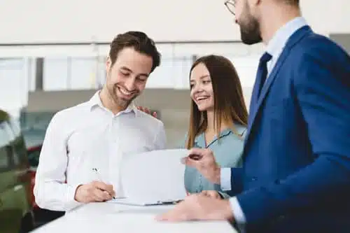 A man in a white shirt happily signs a document while a woman beside him smiles. Another man in a blue suit hands over the paper across a desk. They are indoors, possibly in an office or dealership setting.