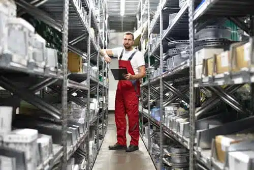 A man in red overalls stands in a warehouse aisle, holding a black folder. Shelves are filled with various boxes and industrial items. The setting is well-lit, and he appears to be conducting an inventory check.