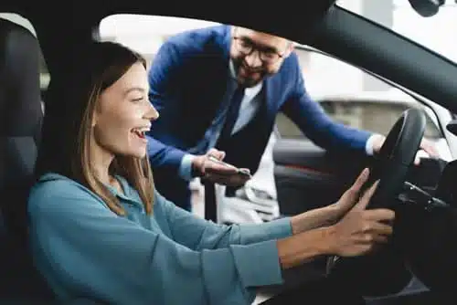 A woman sits in a car, smiling and holding the steering wheel, while a man in a suit leans in through the open window, talking to her. They appear to be in a car dealership or showroom.