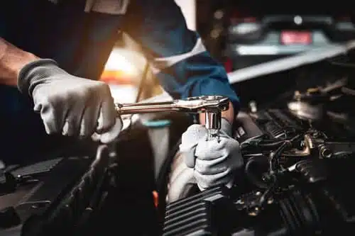 A mechanic wearing gloves uses a ratchet wrench to work on a car engine. The scene is taken up close, showing the detailed parts of the engine and the mechanics focused action. The background is softly blurred.