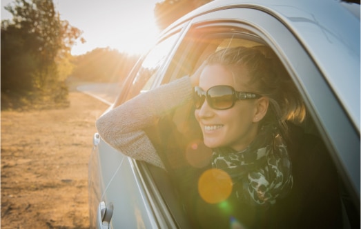 A woman wearing sunglasses and a scarf leans out of a car window, smiling as the sun sets in the background. The bright sunlight creates lens flare, adding a warm glow to the scene. Trees and a road are visible along the cars path.