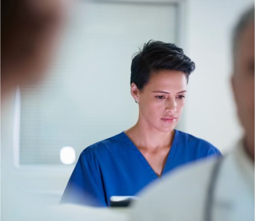 A person in blue medical scrubs stands in a hospital setting, appearing focused and thoughtful. The background is softly blurred, with indistinct figures and a window, suggesting a busy medical environment.