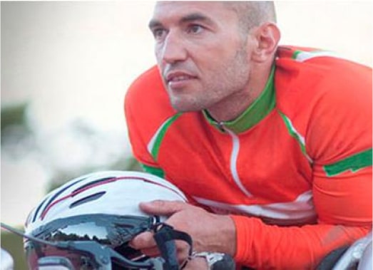 A cyclist in an orange and green outfit holds a helmet while looking into the distance.