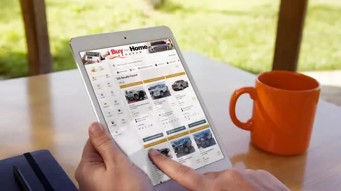 A person is using a tablet to browse a website featuring various cars. The screen displays car listings with images and details. An orange mug and a notebook are on the wooden table, and greenery is visible in the background.