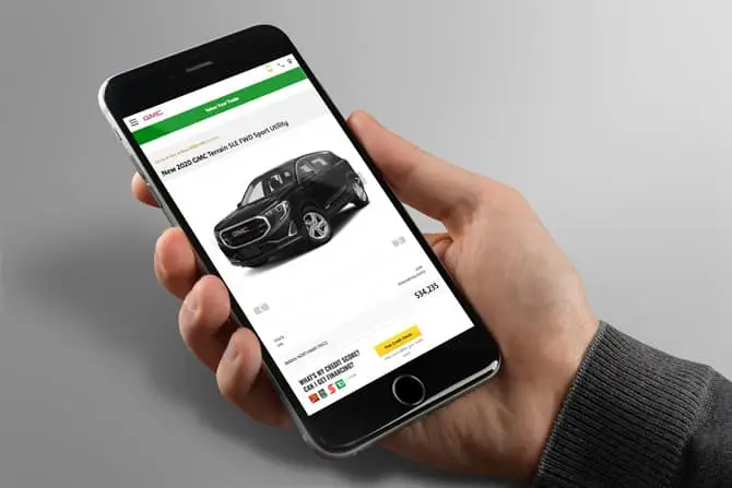 A person holds a smartphone displaying a car sales website. The screen shows a black 2023 GMC Terrain SLE with a price of $31,835. Options to calculate payments and view details are visible. The background is a plain gray surface.