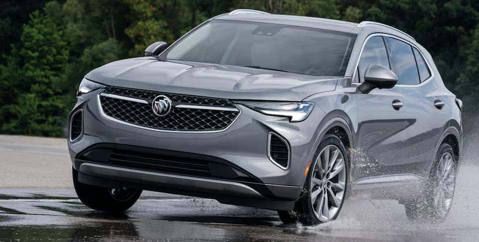 A silver SUV drives through a wet road, creating splashes of water. The vehicle, captured at an angle, showcases its sleek design and modern grille. Trees are visible in the blurred background.