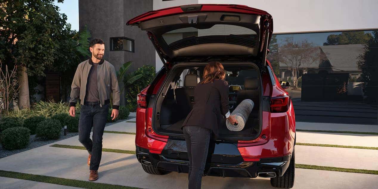 A man and woman are interacting with a red SUV. The woman is placing a rolled-up carpet into the open trunk. The man stands nearby, smiling. They are outdoors on a driveway with greenery and a building in the background.