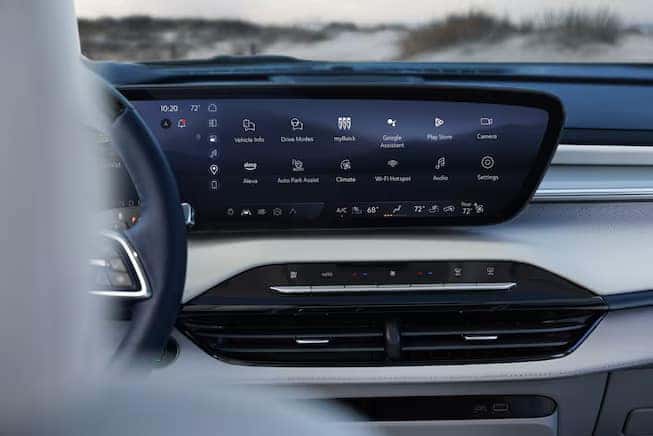 The image shows a car dashboard with a large digital display. The screen shows various options, including vehicle info, drive modes, navigation, and climate control. Below the screen are air vents and control buttons. The scene is outdoors.
