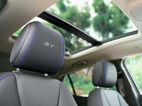 View of a car interior featuring black leather seats with blue stitching, embroidered with ST on the headrest. The sunroof is open, and trees are visible through it, creating a bright and airy atmosphere.