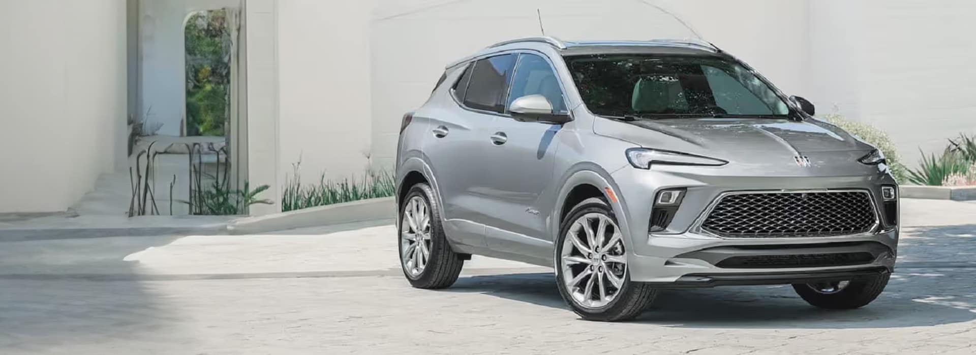 Silver SUV parked on a light-colored driveway in front of a modern building with large windows and plants. The vehicles design features sleek lines and a prominent grille. Bright daylight enhances the cars metallic finish.
