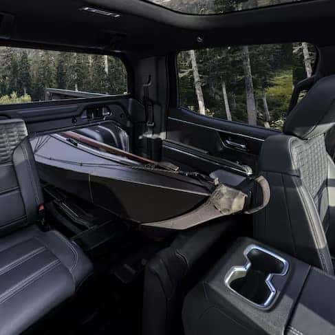 The interior of a vehicle with a spacious backseat featuring folded seats to accommodate a large and sleek cargo item. The window view shows a forested area, and the design includes black leather upholstery and modern amenities.