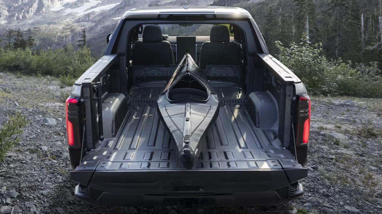 A black kayak is securely placed in the bed of a pickup truck. The truck is parked on a gravel path surrounded by greenery and pine trees, with mountains visible in the background under a clear sky.