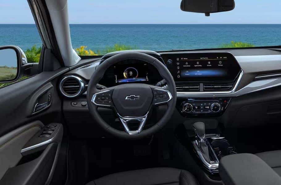 The image shows the interior of a Chevrolet vehicle, focusing on the steering wheel and dashboard. The dashboard displays a digital screen, and the window view shows a clear blue ocean and sky.