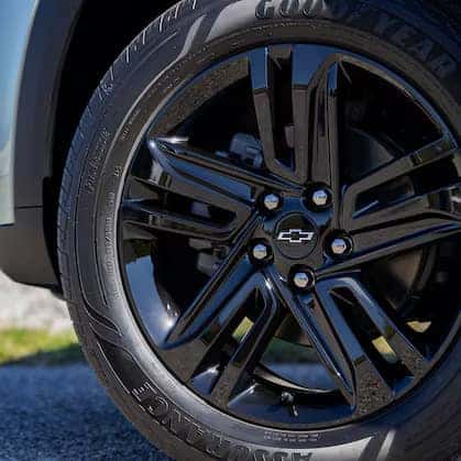 Close-up of a vehicles shiny black alloy wheel, featuring a Chevrolet logo at the center. The tire has clear Goodyear branding. The background is slightly blurred, hinting at a sunny day outdoors.