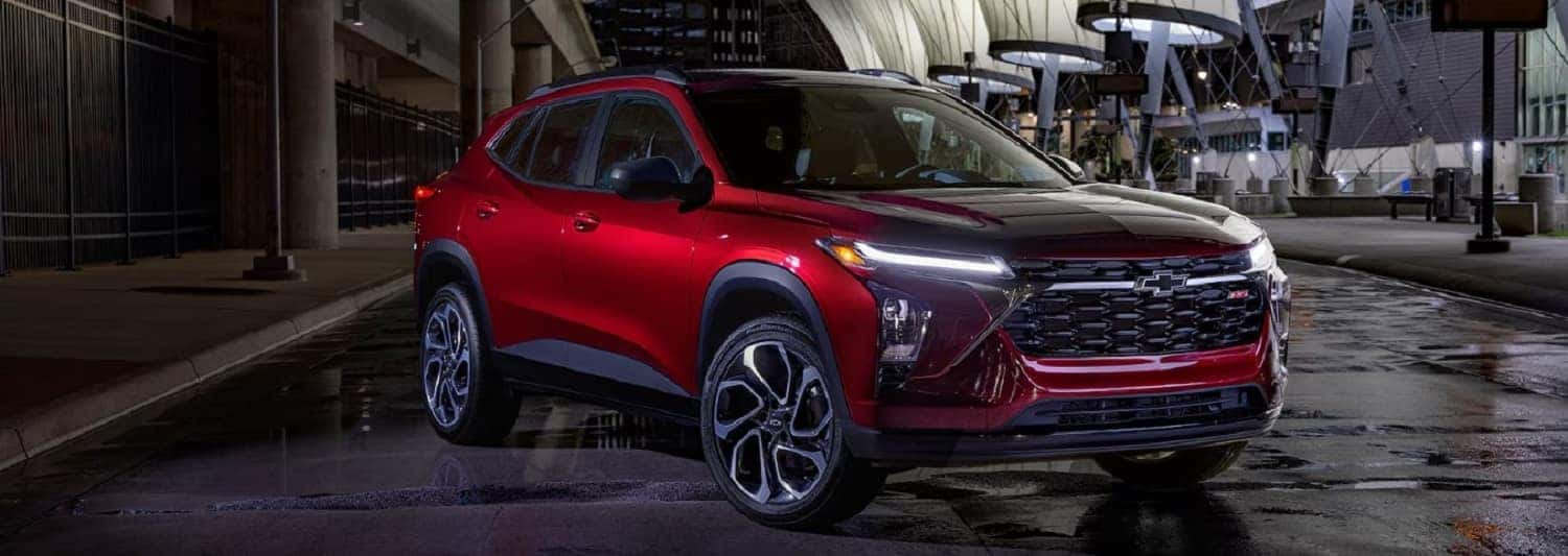 A shiny red SUV is parked in a dimly lit urban setting at night. The car features a sleek design with distinctive wheel rims, and illuminated headlights, reflecting light on the wet pavement. Modern architectural elements are visible in the background.