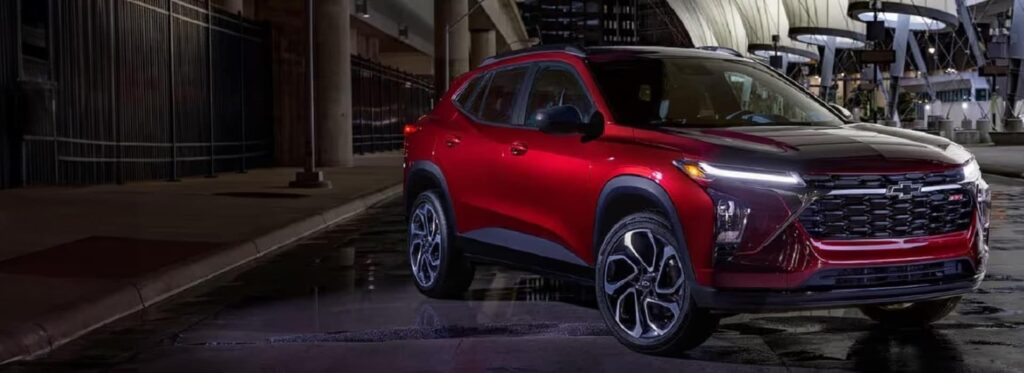 A sleek, red SUV is parked on a wet urban street at night. The vehicles headlights are on, highlighting its modern design and prominent grille. The backdrop features blurred buildings and streetlights, creating a dynamic city atmosphere.