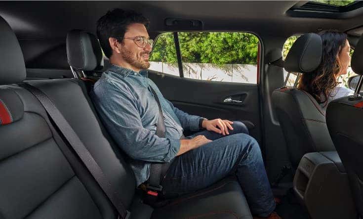 A man wearing glasses and a denim shirt sits comfortably in the back seat of a car, with a woman in the front passenger seat. The car interior is dark with red accents, and trees are visible through the window.