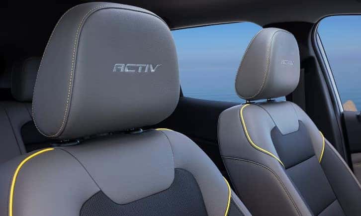 Front seats of a vehicle interior with ACTIV embroidered on the headrests. The seats are gray with yellow stitching. A blurred view of the outside is visible through the window.