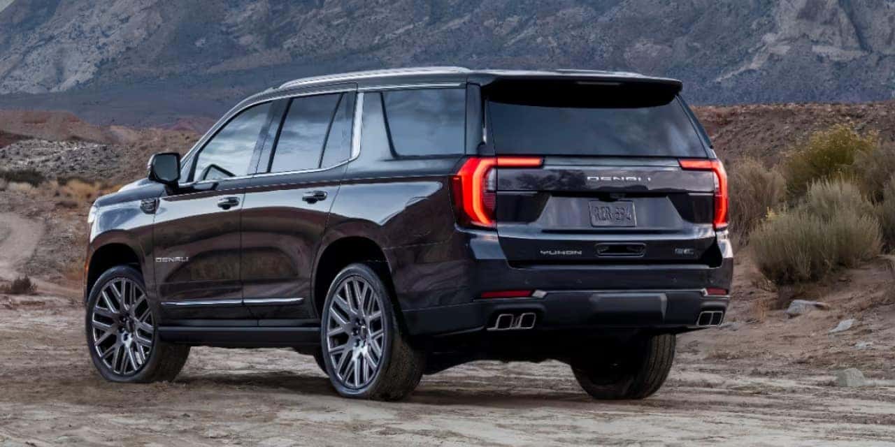 A black SUV parked on a dirt road with mountains in the background. The vehicle has sleek lines, tinted windows, and distinctive taillights. Its positioned at an angle showing the rear and part of the passenger side.