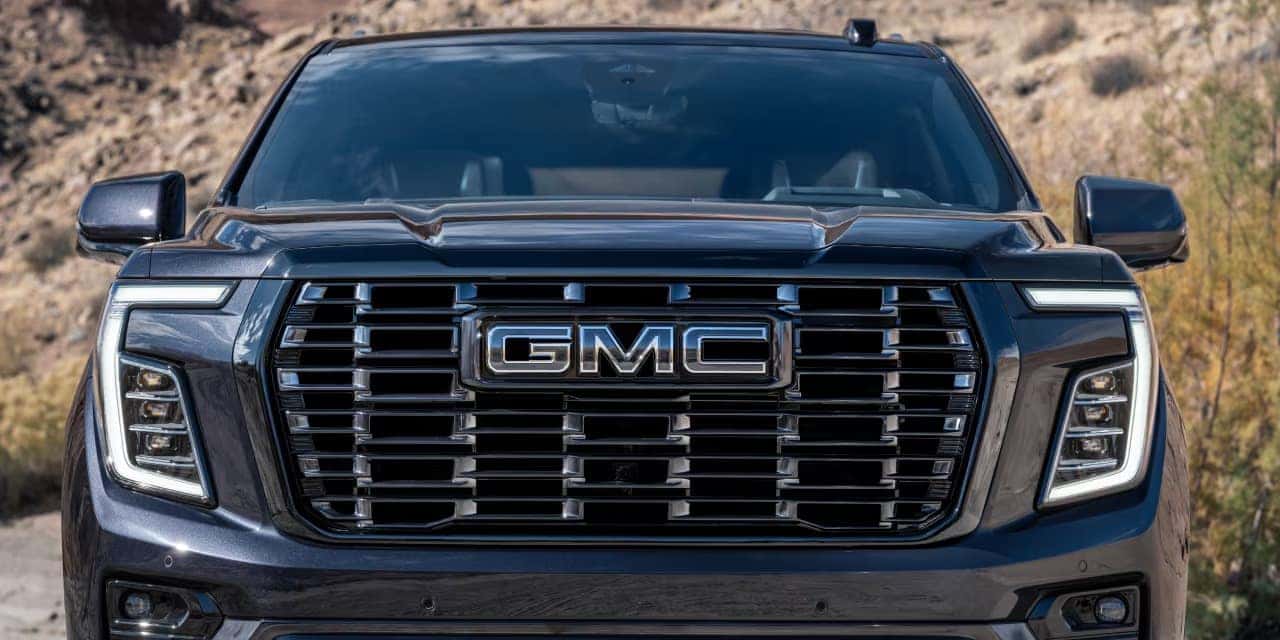 Close-up of a black GMC vehicles front, showcasing the prominent grille with the GMC logo at the center. The SUVs headlamps are on either side, set against an outdoor background with dry, grassy terrain and a rocky area.