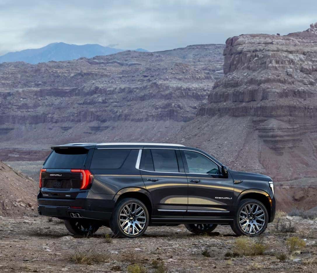 A black SUV is parked on a rocky terrain with a background of vast, rugged canyons and distant mountains under a cloudy sky. The vehicle is facing right, highlighting its side and rear.