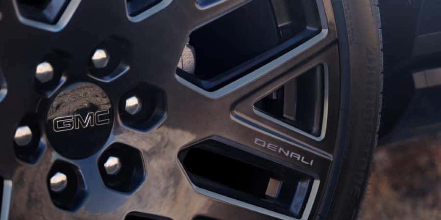 Close-up of a GMC Denali wheel, featuring a modern, geometric alloy design. The center cap displays the GMC logo, and Denali is engraved on the spoke. The wheel is mounted on a black tire with visible treads.