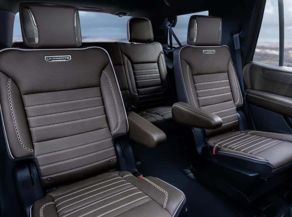 Luxury vehicle interior with three rows of brown leather seats. The seats have Ultimate labels, prominent stitching, and headrests. The background shows a blurred outdoor landscape through large windows.