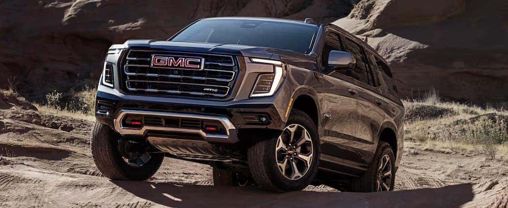 A black GMC SUV navigates a rugged, sandy terrain. The vehicles grille and headlights are prominently visible, and its wheels are gripping the uneven ground. The background features rocky formations under a clear sky.