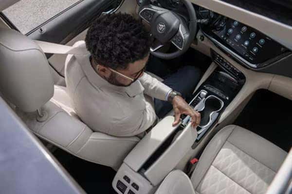 Man with glasses sitting in the drivers seat of a car, viewed from above. He is reaching toward the center console storage area. The car interior features a touchscreen dashboard and beige leather seats.