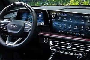 Interior view of a car featuring a sleek dashboard and steering wheel. The dashboard displays multiple digital screens with icons and controls. The steering wheel has a recognizable logo in the center. Sunlight filters in from the windows.
