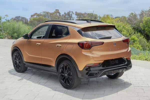 A bronze crossover SUV is parked outdoors on a stone driveway. The vehicle has black accents and wheels. Lush greenery and trees are visible in the background.