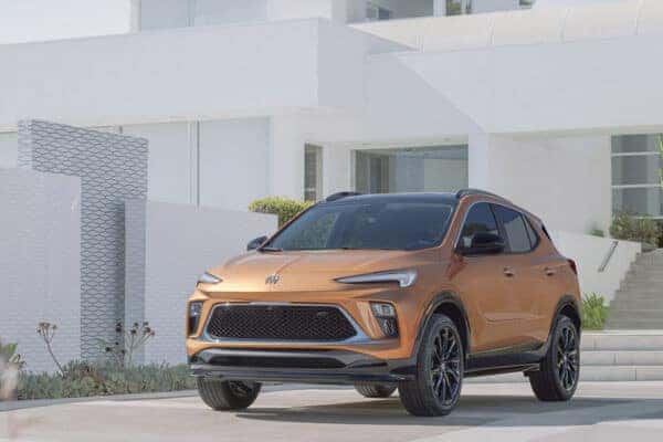A bronze-colored SUV is parked outside a modern, white building with large windows. The vehicle has a sleek design with black accents and is surrounded by minimalistic landscaping.