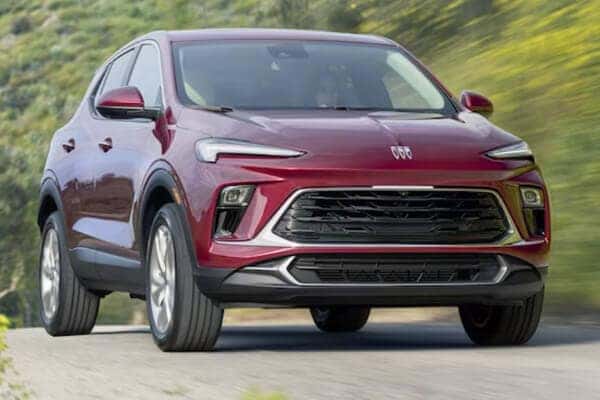 A red SUV is driving on a road with greenery in the background. The vehicle has a sleek design with prominent headlights and a large front grille. The scene suggests motion and a natural outdoor setting.