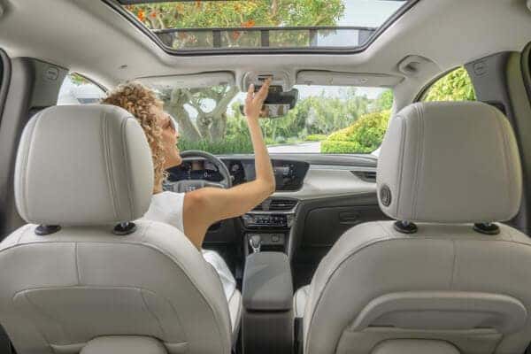 A person with curly hair sits in the drivers seat of a car, reaching up to adjust the rearview mirror. The cars interior is equipped with a sunroof, and lush greenery is visible outside the windows.