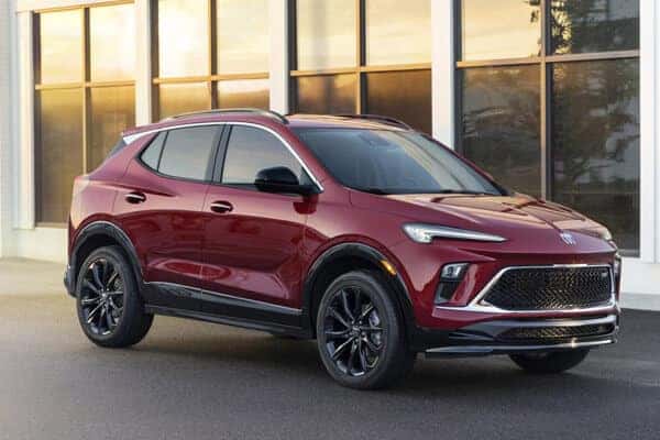 A red SUV is parked in front of a building with large windows. The vehicle has black wheels and a sleek, modern design, reflecting light from its glossy surface. The background shows a warm, sunset glow.
