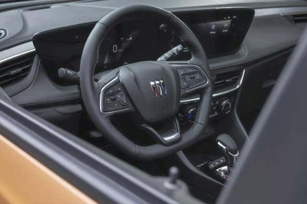 Interior view of a car showing a black steering wheel with a Buick logo, dashboard with digital display, and gear shift. The dashboard controls include buttons and knobs. The car has a modern and sleek design.