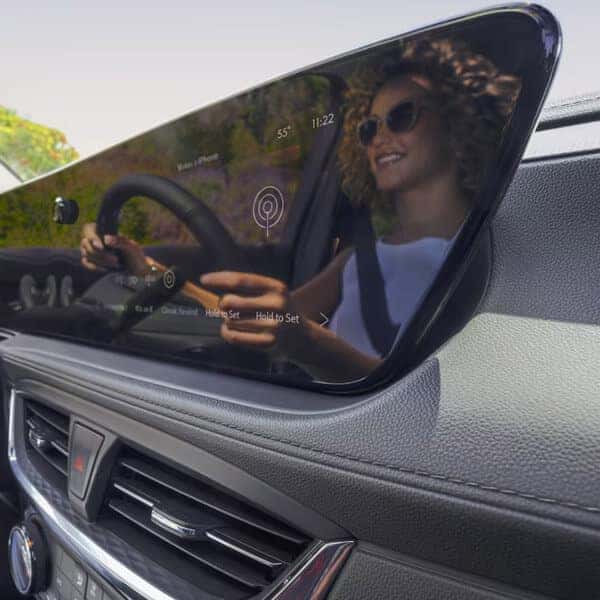 A woman with curly hair and sunglasses drives a car, reflected in a large digital display on the dashboard. The display shows icons and text indicating weather and navigation. Sunlight filters through the windshield, highlighting the interior.