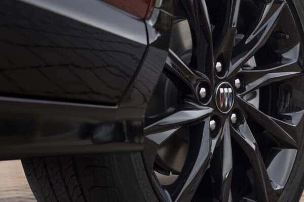 Close-up of a cars black alloy wheel with a visible logo at the center. The wheel features multiple sleek spokes, and the tire is detailed with tread patterns. Part of the car’s bodywork is visible in the background.