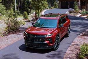 A red SUV is parked on a paved driveway surrounded by lush greenery and trees. A house is visible in the background. The vehicle is shiny and appears to be a modern model.