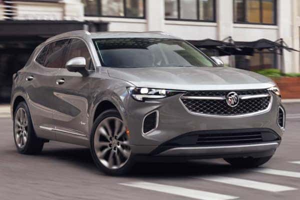 A silver SUV is driving on a city street. The vehicle has a sleek design with a wide front grille and modern headlights. Buildings and a sidewalk with plants are visible in the background.