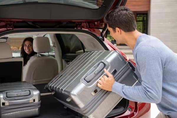 A person loads a large silver suitcase into the trunk of a red SUV. Another person sits in the drivers seat, smiling and looking back at the person with the suitcase. The vehicle is parked outside a building with a wooden facade.