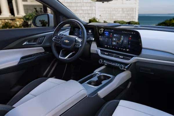 Interior of a car showcasing a modern dashboard with a large touchscreen display, steering wheel, and center console. The seats are upholstered in a combination of black and gray materials. A scenic ocean view is visible through the window.