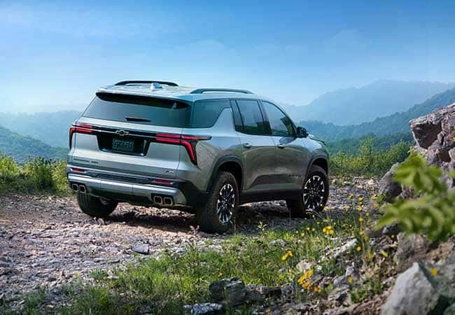 A silver SUV parked on a rocky, mountainous terrain with a scenic view of green hills and a clear blue sky in the background. Yellow flowers are visible among the rocks in the foreground.