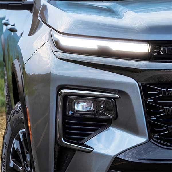 Close-up of the front left side of a dark gray truck, focusing on the sleek modern headlight design. The image shows the angular lines and LED lights against a blurred natural background.